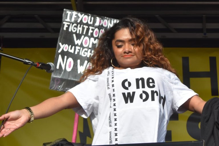 Chedda is standing on stage with a white t-shirt with the new OneWorld-logo on it. The logo are black letters. Her arms are spread to show the crowd.